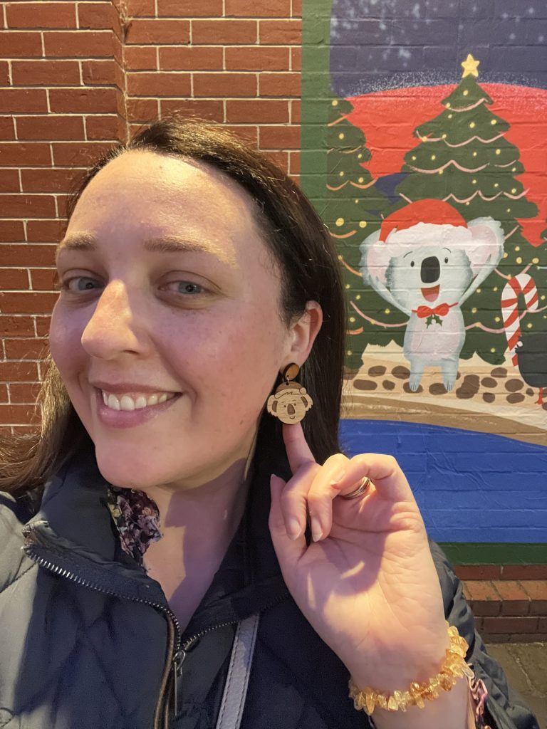 Liv Lorkin wearing a koala Christmas earring in front of a mural of a scene from her Christmas in Ballarat children's book, which is part of the 2022 Christmas art trial. The mural features a colorful depiction of the magical city of Ballarat.