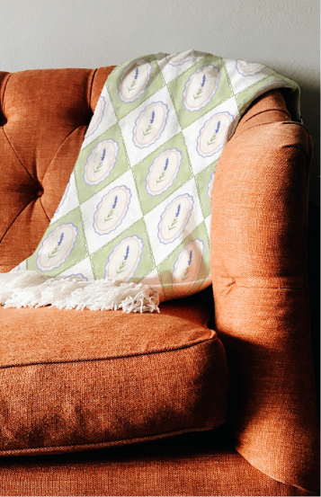 Watercolour illustration by Liv Lorkin featuring a stunning diamond pattern blanket with alternating designs of intricate purple flowers with green leaves, one against a green background, all set against a cozy brown couch.